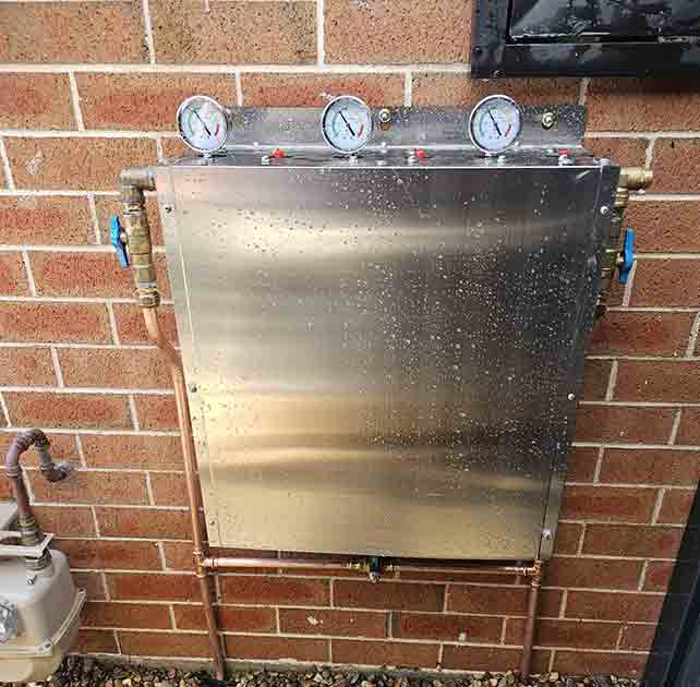 Hot water system repair unit mounted on a brick wall with pressure gauges and plumbing connections, representing GPS Plumbing Services' expertise in hot water system maintenance and repair in Sydney.